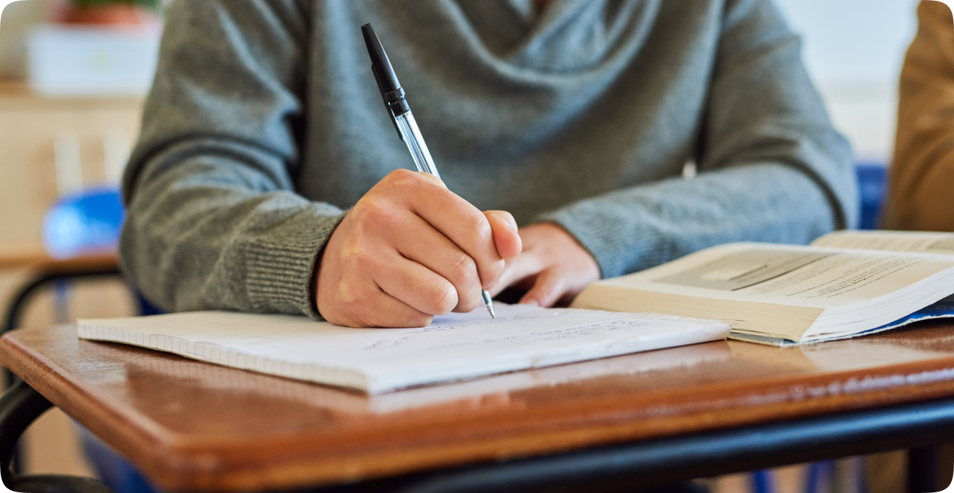 Someone sat at a desk writing in their notebook while reading a textbook.